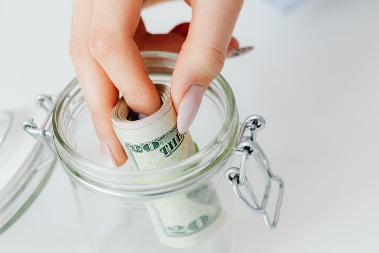 Un primer plano de una mano colocando dólares enrollados en un frasco de vidrio, simbolizando el ahorro, recursos y educación financiera.