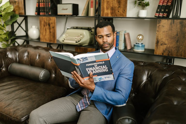 Un hombre con una chaqueta azul está sentado en un sofá de cuero leyendo un libro de los 17 archivos de riqueza.
