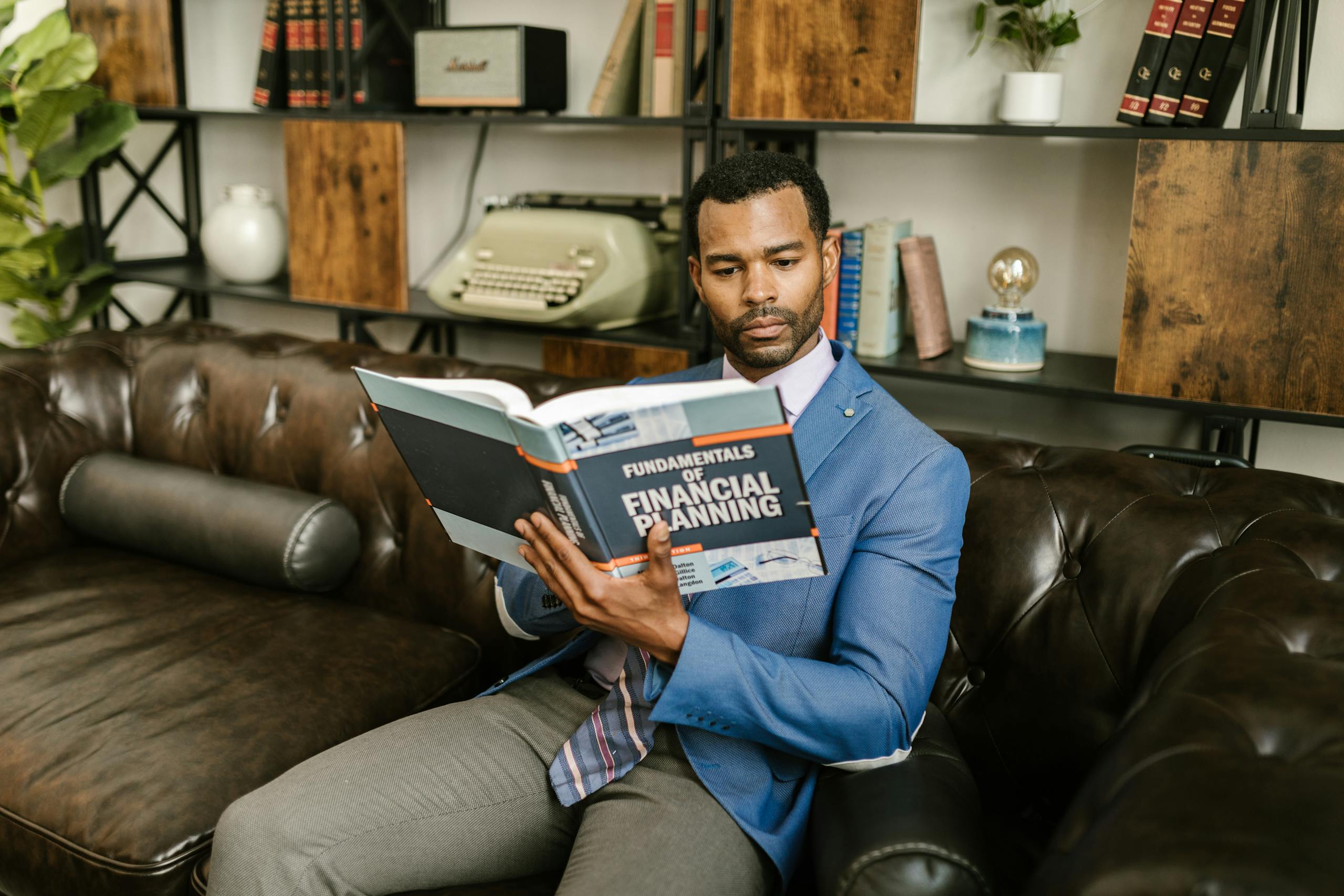 A man in a blue blazer sits on a leather couch reading a financial planning book.