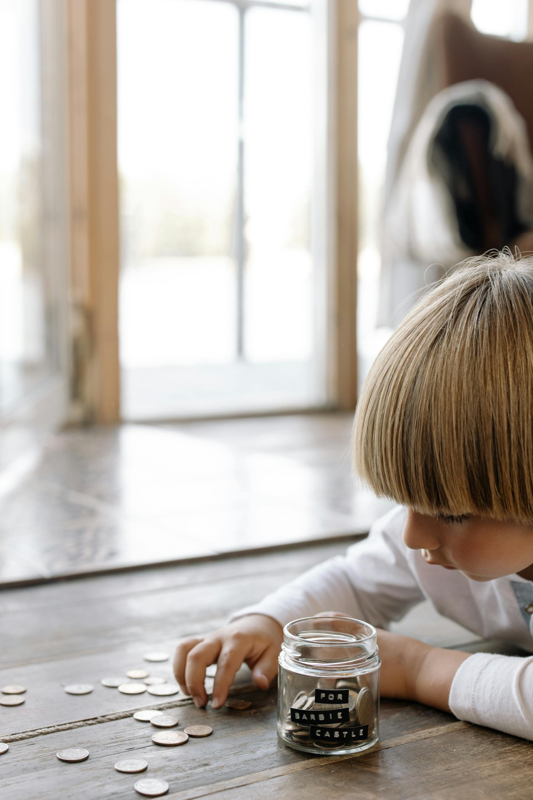 Un niño pequeño contando monedas y colocándolas en un frasco etiquetado "para el castillo de Barbie" en el interior de un piso de madera simbolizando educación financiera desde niños.