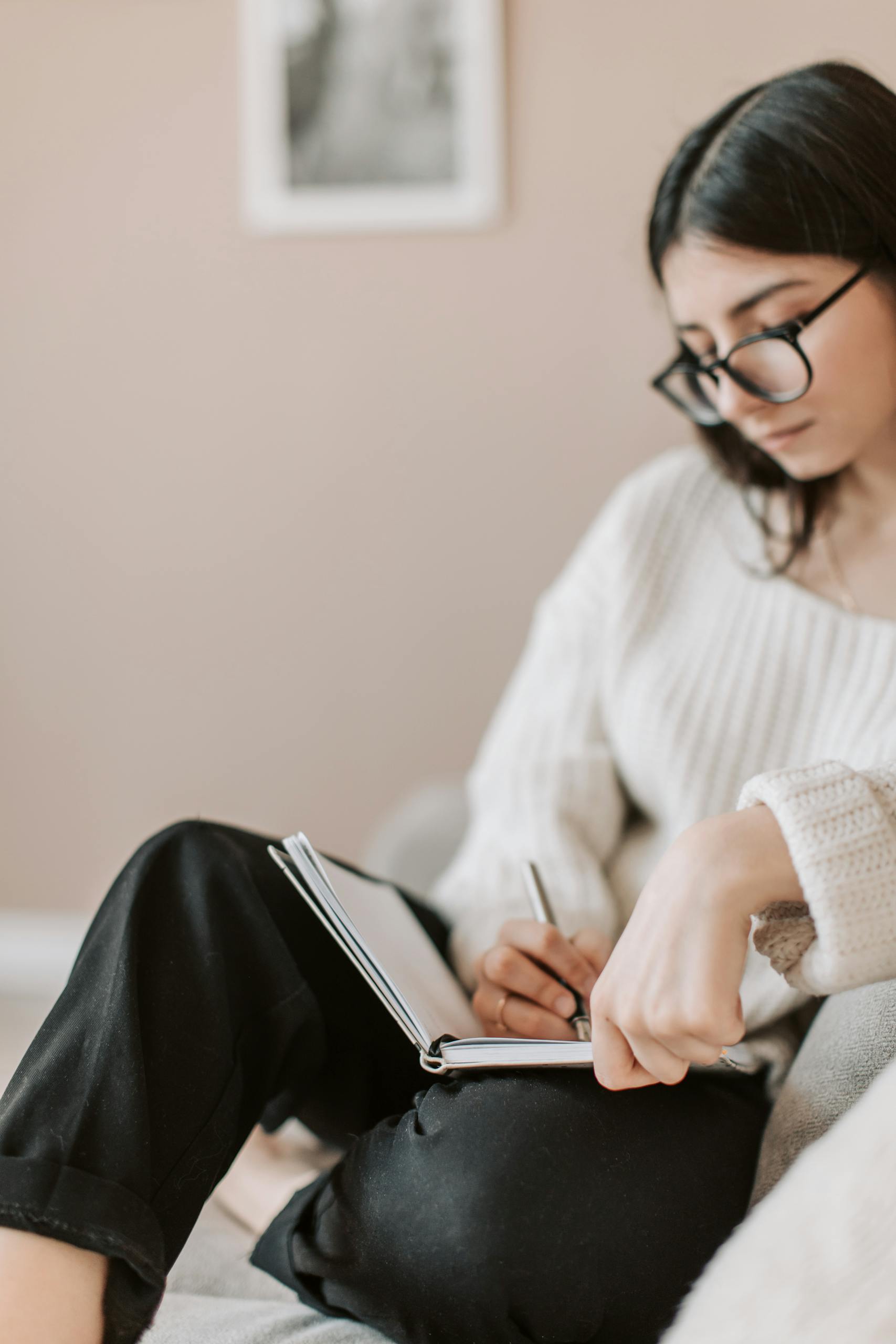 Mujer atenta con gafas y ropa informal sentada descalza con las piernas cruzadas en un cómodo sofá en un piso moderno y tomando notas en un cuaderno con bolígrafo