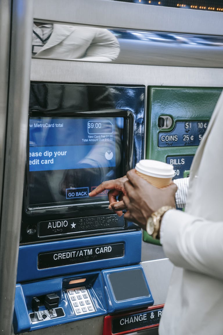 Close-up of a person using a subway ticket machine with a credit card and coffee cup.