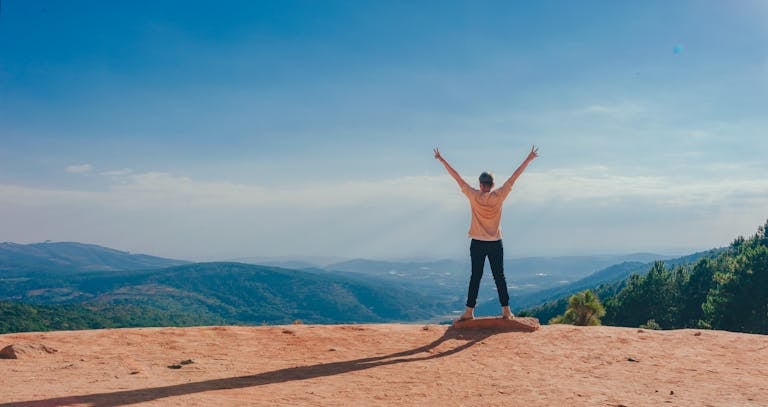 Un hombre levanta los brazos en señal de triunfo en la cima de una montaña rocosa con vistas a un vasto paisaje simbolizando la creación de su futuro millonario.