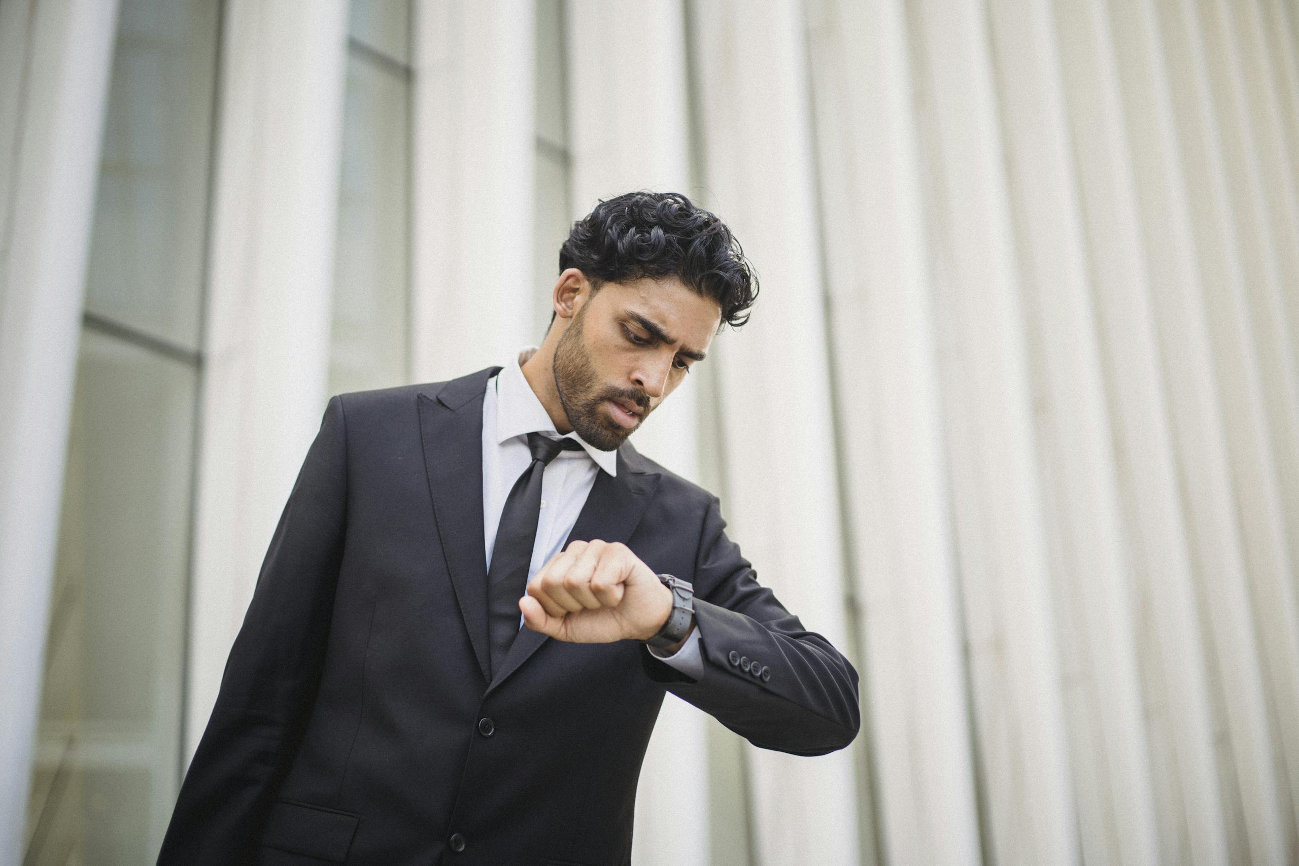 Un hombre con traje negro mira su reloj frente a un edificio moderno, representando puntualidad en el cumplimiento de pagos.